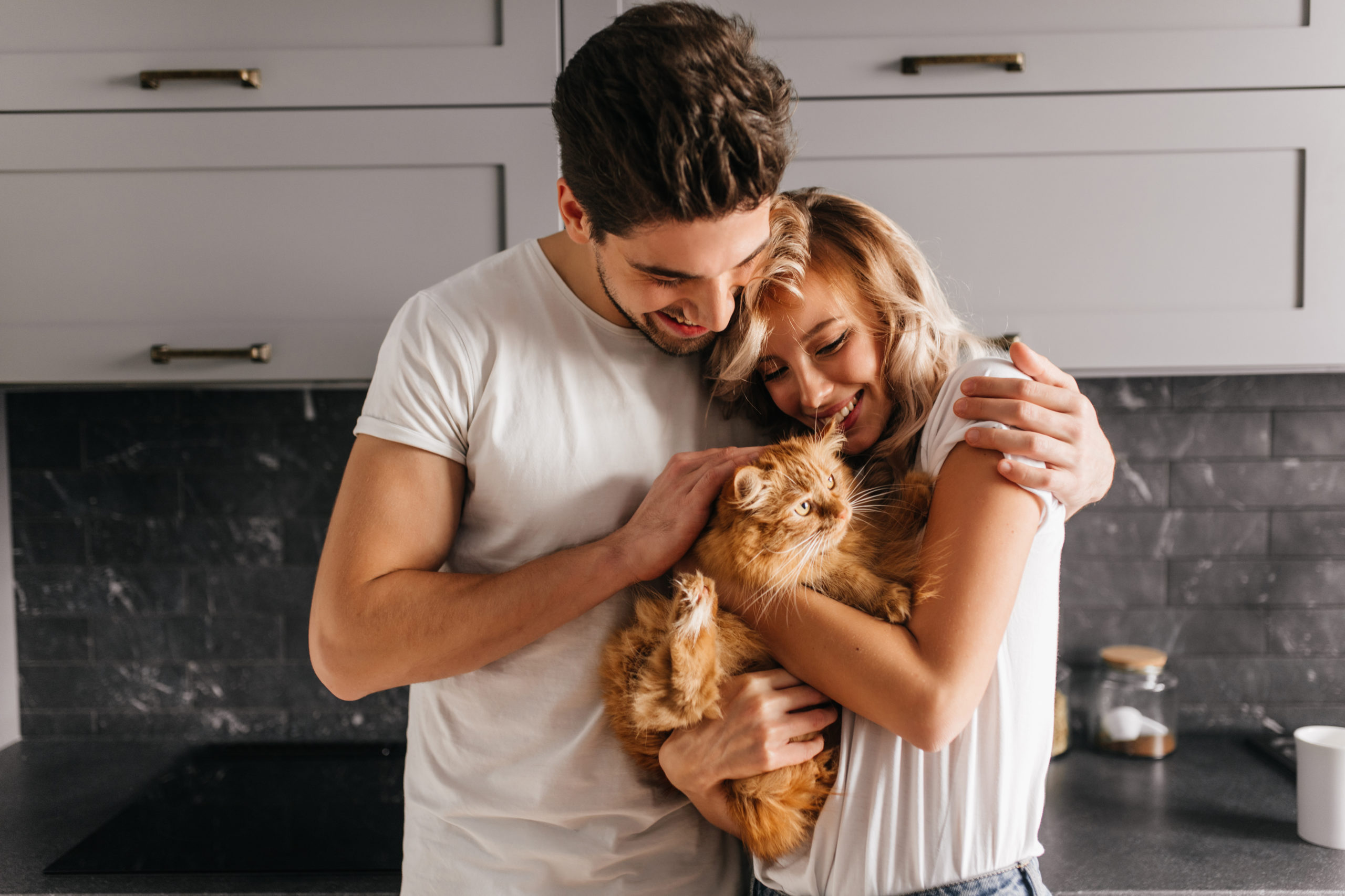 Brunette man looking at his cat and embracing wife. Indoor photo of happy family posing with pet
